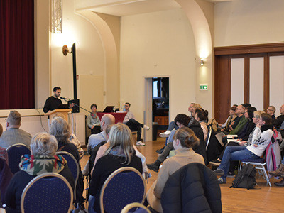 Zuschauerperspektive: Gäste von hinten und von der Seite fotografiert blicken zum Stehpult, an Maher Seger vom KSL.Münster referiert. Der Raum im Martin-Luther-Haus in Steinfurt wirkt groß, der Boden besteht aus braunem Parkett und es ist ein roter Samtvorhang im Hintergrund zu sehen.