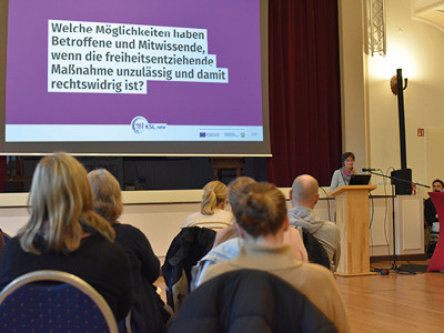 Zuschauerperspektive: Gäste von hinten fotografiert blicken nach vorne zum Redebeitrag von Martina Steinke vom KSL.Münster, die an einem Stehpult rechts neben einer Leinwand mit Präsentation steht. Auf der Präsentation steht die Frage: Welche Möglichkeiten haben Betroffene und Mitwissende, wenn die freiheitsentziehende Maßnahme unzulässig und damit rechtswidrig ist?