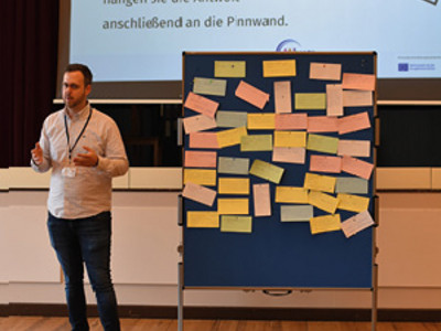 Timo Krasemann – Männerberater für Menschen mit Behinderungen und Deeskalationstrainer im Familienunterstützenden Dienst Steinfurt – steht vor der Pinnwand mit den Informationen, die die Teilnehmenden darauf hinterlassen haben. Hinter ihm ist seine Präsentation auf einer Leinwand zu sehen. Darauf steht: Welches Beispiel von Gewalt ist Ihnen auf der Arbeit begegnet? Notieren Sie eine Antwort auf einen Zettel. Wenn Sie möchten, hängen Sie die Antwort auf die Pinnwand.