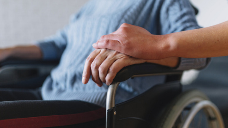 Frau im Rollstuhl, ihre linke Hand wird von Hand einer Besucherin gehalten